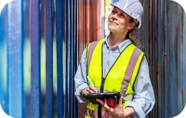 Factory worker in safety vest inspecting stacked materials with a handheld device.
