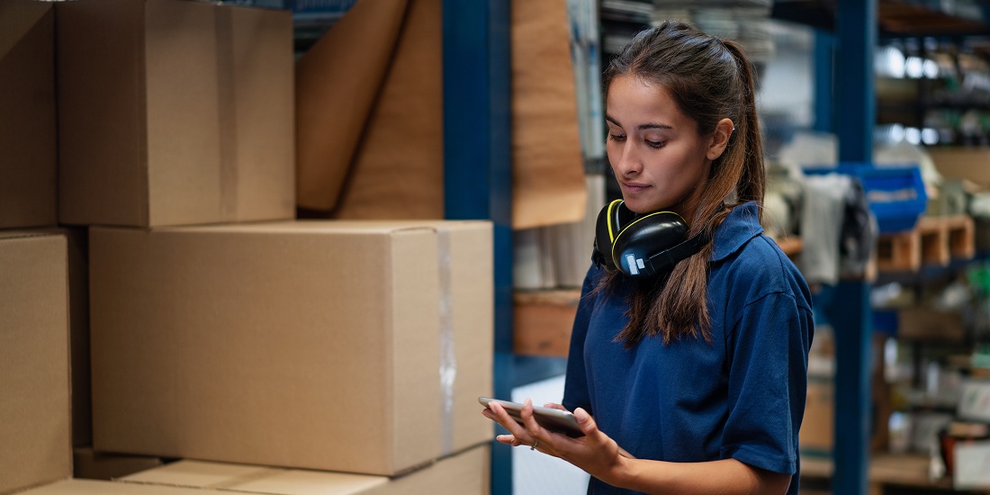 Supply chain specialist checking inventory on tablet in warehouse surrounded by boxes.
